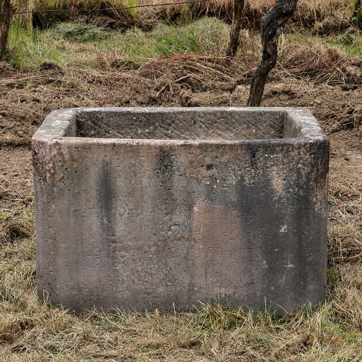 Ein jahrhundertealter Sandsteinkübel (75 x 53 x 48), rechteckig und verwittert, steht auf trockenem Gras, im Hintergrund sind ein Feld und einige Pflanzen zu sehen.