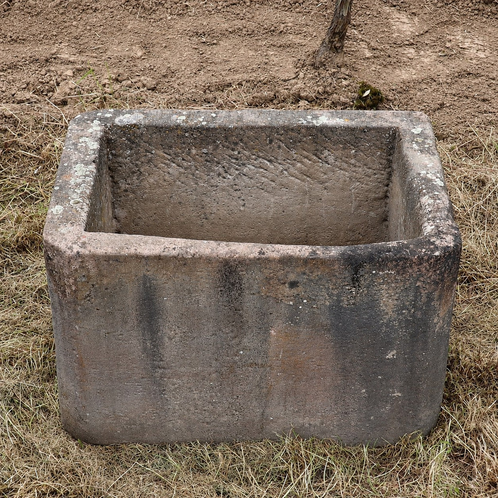 Ein großer, rechteckiger Behälter aus verwittertem Beton steht auf trockenem Gras und Erde; seine raue Oberfläche und die dunklen Flecken erinnern an einen jahrhundertealten Sandsteinkübel (75 x 53 x 48), wie man ihn vielleicht findet, wenn man nach „historischem Sandsteinkübel kaufen“ sucht.