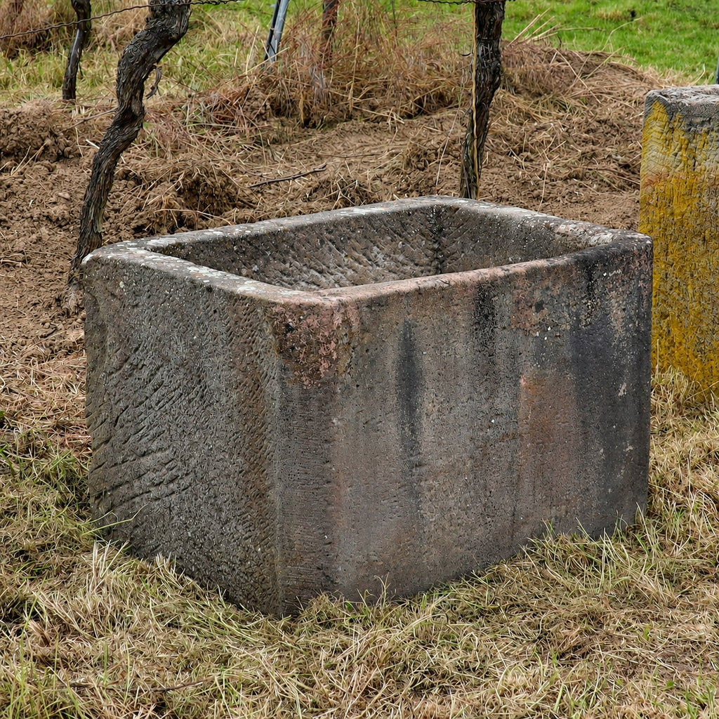 Ein großer, rechteckiger, verwitterter Steinkübel steht auf trockenem Gras, umgeben von spärlicher Vegetation und Holzpfosten – eine ideale Inspiration, wenn Sie auf der Suche nach einem jahrhundertealten Sandsteinkübel (75 x 53 x 48) für Ihren eigenen rustikalen Außenbereich sind.