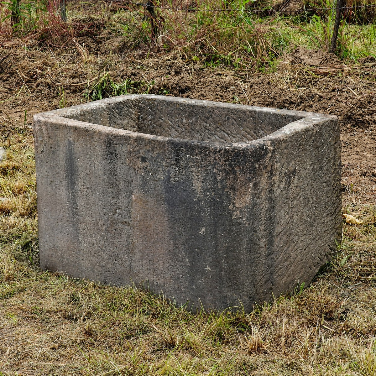 Ein großer, rechteckiger Sandsteinkübel aus dem 19. Jahrhundert (75 x 53 x 48) steht auf einer Rasenfläche in der Nähe eines Drahtzauns, im Hintergrund sind Erde und Unkraut zu sehen – eine ideale Wahl für alle, die einen historischen Sandsteinkübel kaufen möchten, um ihren Außenbereich mit zeitlosem Charme zu bereichern.