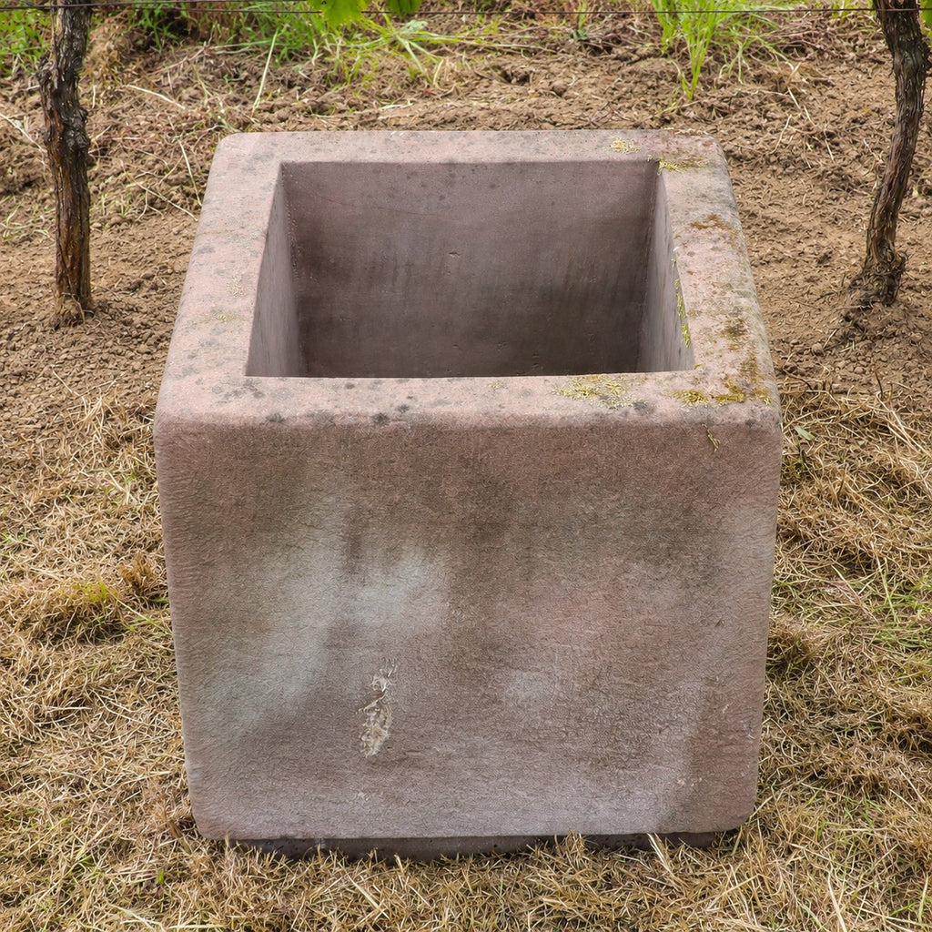 Auf trockenem Gras steht ein großer, leerer, quadratischer Betonpflanzkübel, im Hintergrund sind etwas Erde und grüne Pflanzen zu sehen – ideal für alle, die ein 2er-Set Natursteintröge 60 x 60 x 56 kaufen und ihrem Garten zeitlosen Charme verleihen möchten.