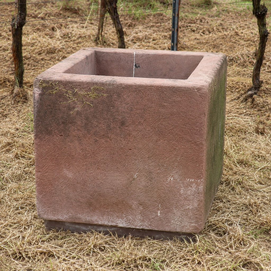 Ein großes, quadratisches 2er-Set Natursteintröge (60 x 60 x 56) steht auf trockenem Gras, im Hintergrund sind einige Ranken und ein Drahtzaun zu sehen. Wenn Sie Ihrem Garten zeitlosen Charme verleihen möchten, sollten Sie diesen Pflanzkübel in Betracht ziehen – kaufen Sie das 2er-Set Natursteintröge 60 x 60 x 56 als eleganten Blickfang für Ihren Außenbereich.