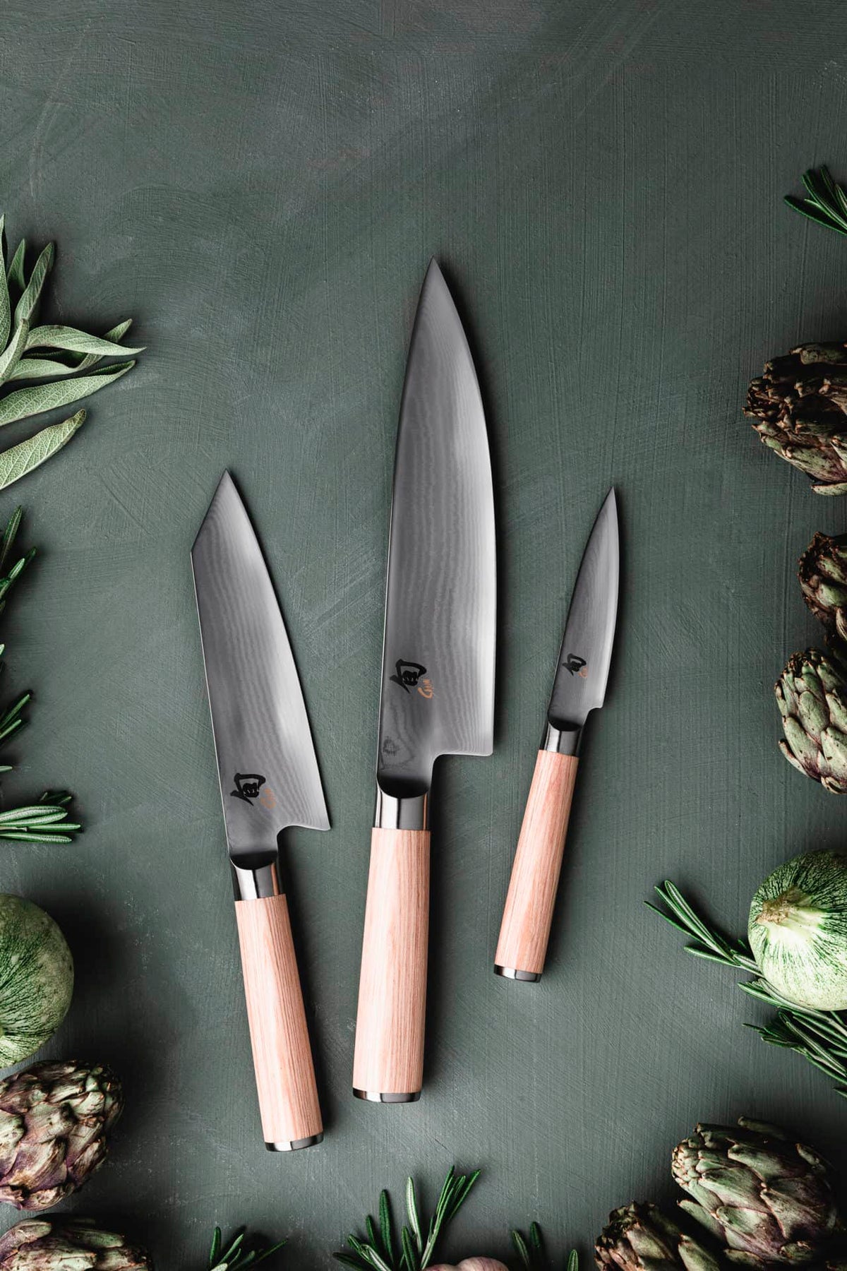 Ein stilvolles Arrangement besteht aus einem Set von drei Messern mit Holzgriffen, wobei ein Kai Shun Classic White Kiritsuke DM-0777W Kochmesser in limitierter Auflage (5555 Stück) prominent in Szene gesetzt wird, ergänzt durch ein großes Kochmesser, ein mittleres Allzweckmesser und ein kleines Schälmesser auf einer grünen Oberfläche. Artischocken und Rosmarinzweige schmücken die Szene kunstvoll.