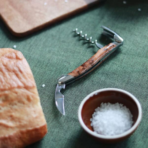 Ein Forge De Laguiole Kellnermesser Sommelier mit mattiertem Wacholder-Griff liegt auf einer grünen Tischdecke neben einem Laib Brot und einer kleinen Schale mit grobem Salz. Im Hintergrund rundet ein hölzernes Schneidebrett den idealen Rahmen für die Freuden eines jeden Sommeliers ab.