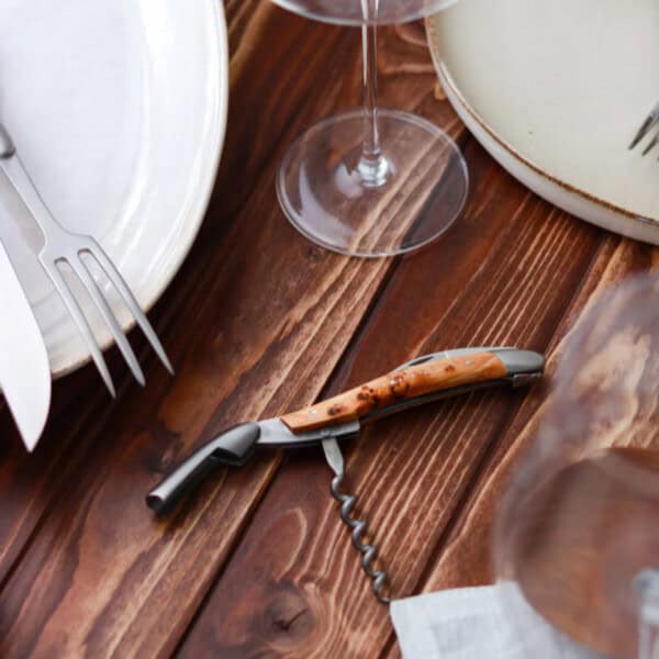 Ein hölzernes Tischgedeck mit weißen Tellern, Gabeln und einem Weinglas präsentiert neben dem Geschirr das charmante Forge De Laguiole Kellnermesser Sommelier im Wacholder-Matt-Finish mit Holzgriff.