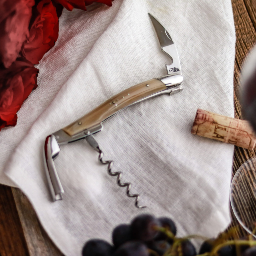 Ein Kellnermesser Sommelier von Forge De Laguiole mit Horngriff ruht auf einem weißen Tuch und verkörpert raffinierte Eleganz. Ein Weinkorken, rote Blumen und Trauben sind kunstvoll in der Nähe auf einer Holzoberfläche arrangiert.