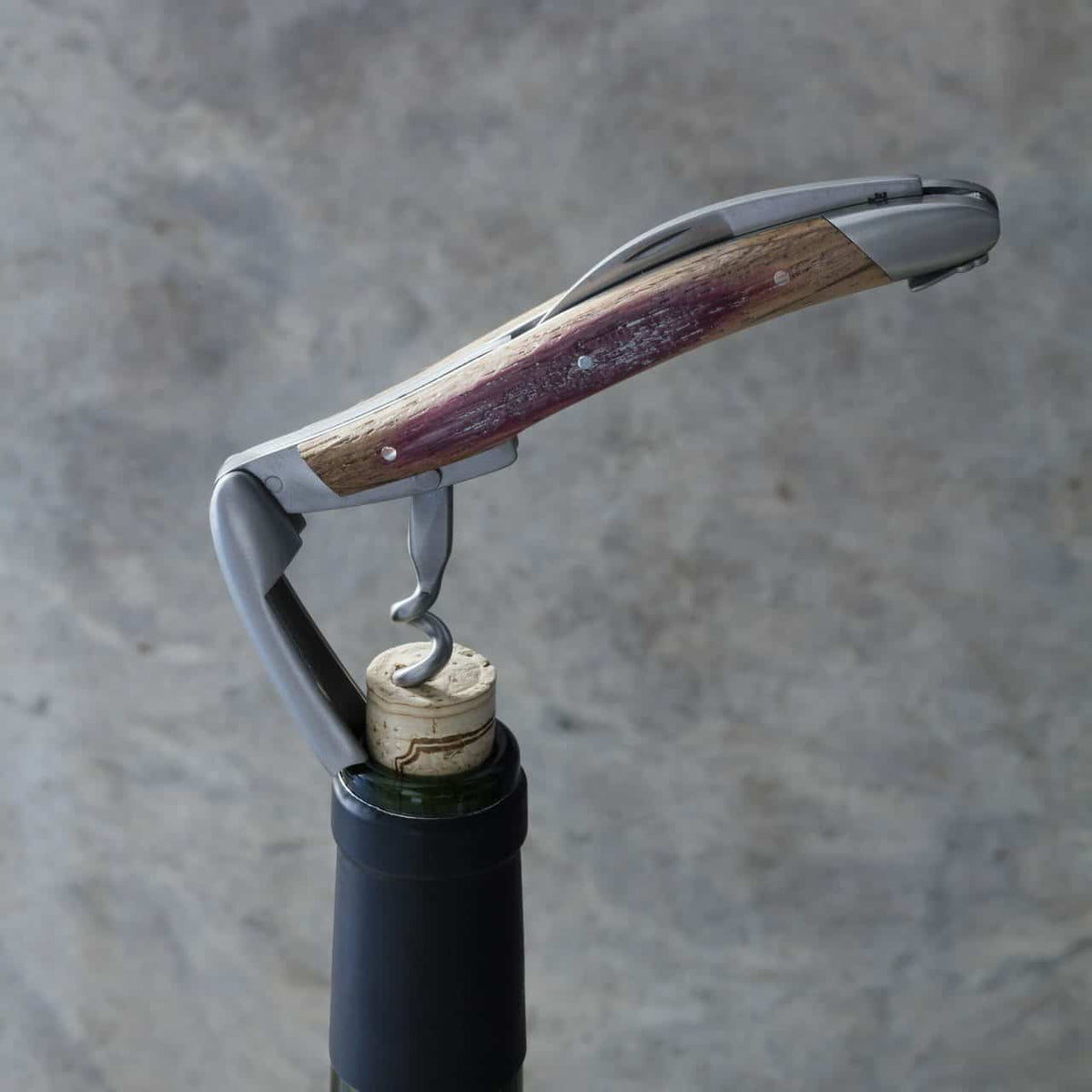 Eine Nahaufnahme eines Korkenziehers, der zum Öffnen einer Weinflasche verwendet wird. Der Korken ist teilweise aus dem Flaschenhals entfernt. Der Korkenzieher hat einen metallischen Hebel und einen Holzgriff mit verwitterter Oberfläche. Der Hintergrund ist eine strukturierte graue Oberfläche.