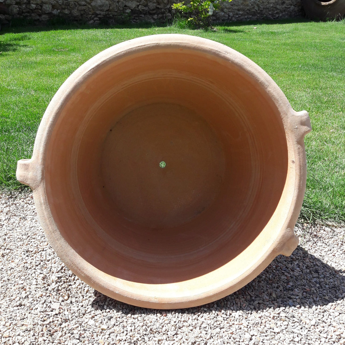 Der Terrakotta-Pflanzkübel „Sorbus“ mit Henkeln (Durchmesser 40–60 cm) ist leer abgebildet, mit einem zentralen Abflussloch, von oben betrachtet, während er auf Kies steht, im Hintergrund grünes Gras und eine Steinmauer.