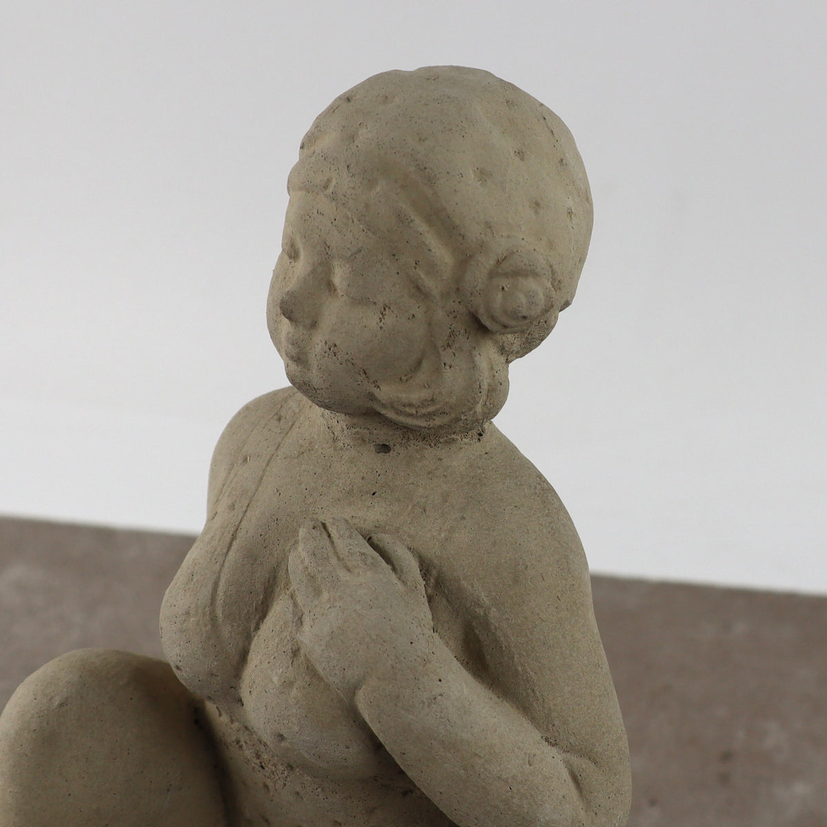 Eine Nahaufnahme der UV-beständigen Gartenfigur "Badenixe" zeigt eine sitzende weibliche Figur mit einer Hand auf der Brust, weichen Gesichtszügen und gewelltem Haar, die sich von einem schlichten Hintergrund abhebt.
