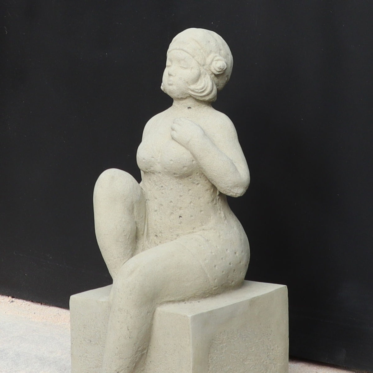 Die UV-beständige Gartenfigur "Badenixe" zeigt eine sitzende Frau im strukturierten Badeanzug und Badekappe, mit erhobenem Knie und Hand auf der Brust - eine stilvolle Gartendekoration vor schwarzem Hintergrund.