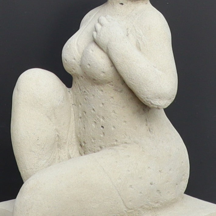 Die UV-beständige Gartenfigur "Badenixe" ist eine Stein-Skulptur einer knienden, nackten Person mit stilisierten, runden Formen und einer Hand auf der Brust - eine auffällige Gartendekoration vor dunklem Hintergrund.