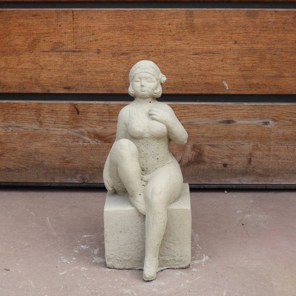 Die UV-beständige Gartenfigur "Badenixe" zeigt eine elegante, sitzende Nixe mit Blume im Haar und übereinandergeschlagenen Beinen auf einem quadratischen Sockel - perfekt als stilvolle Gartendekoration vor Holz.