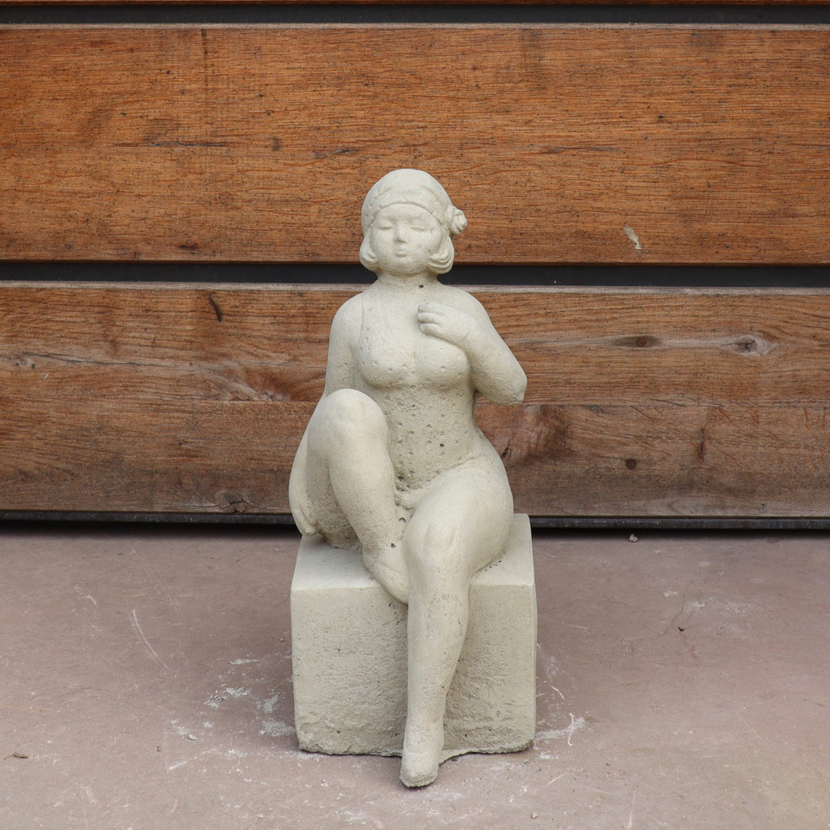 Die UV-beständige Gartenfigur "Badenixe" zeigt eine elegante, sitzende Nixe mit Blume im Haar und übereinandergeschlagenen Beinen auf einem quadratischen Sockel - perfekt als stilvolle Gartendekoration vor Holz.