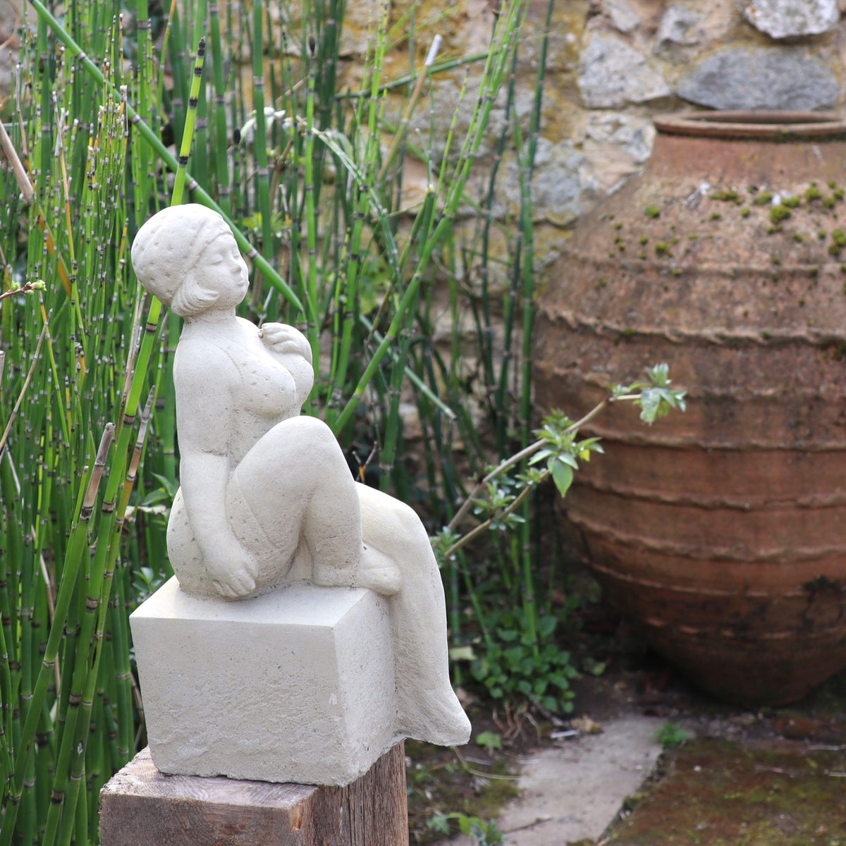 Die UV-beständige Gartenfigur "Badenixe" - eine kleine Statue einer sitzenden Frau mit kurzem Haar - steht als Gartendekoration auf einem Sockel, umgeben von grünen Pflanzen und einem großen, rustikalen Tontopf.