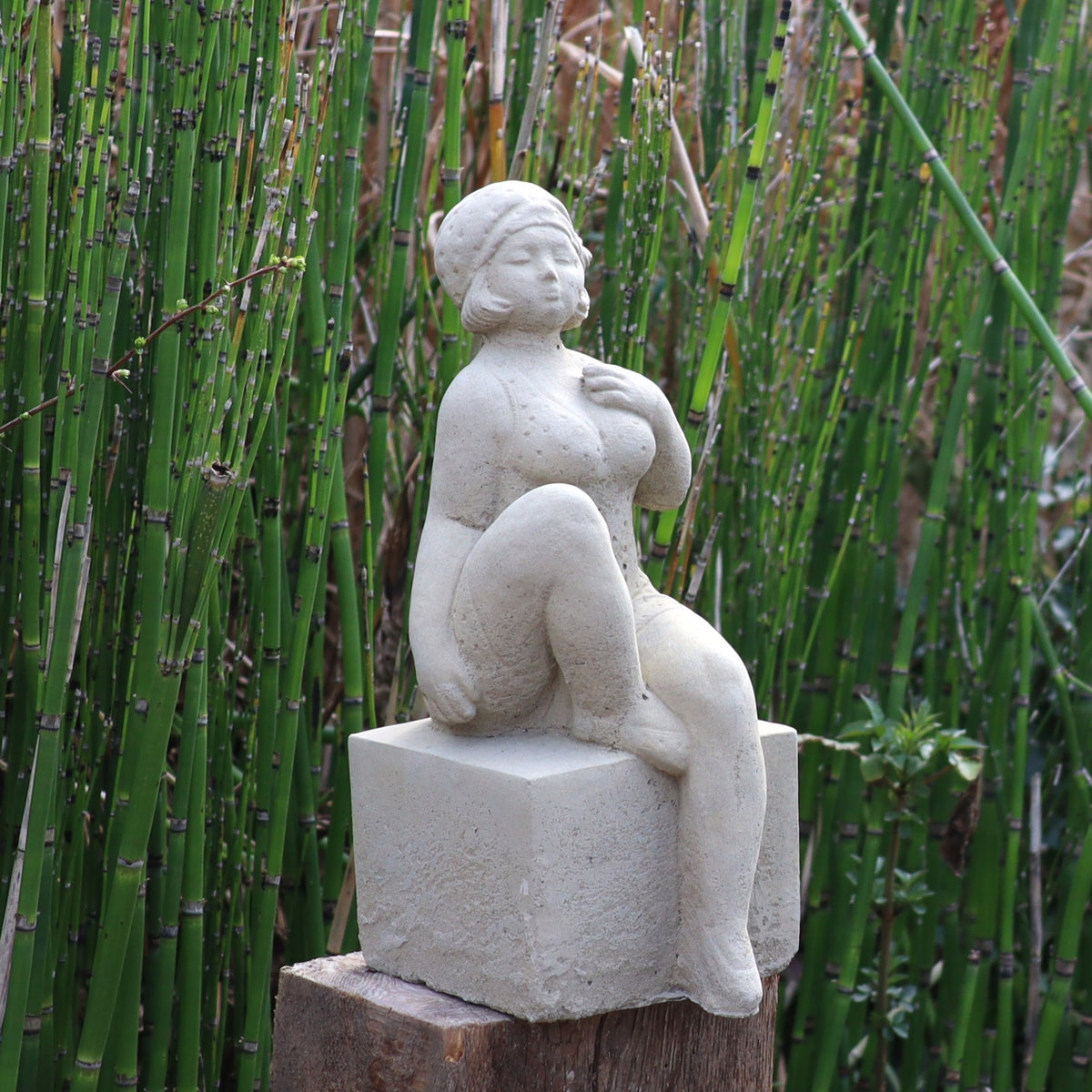 Die UV-beständige Gartenfigur "Badenixe" zeigt eine sitzende, nackte Frau auf Holzsockel und ist von hohen, bambusähnlichen Pflanzen umgeben - eine elegante Dekoration für jeden Garten.