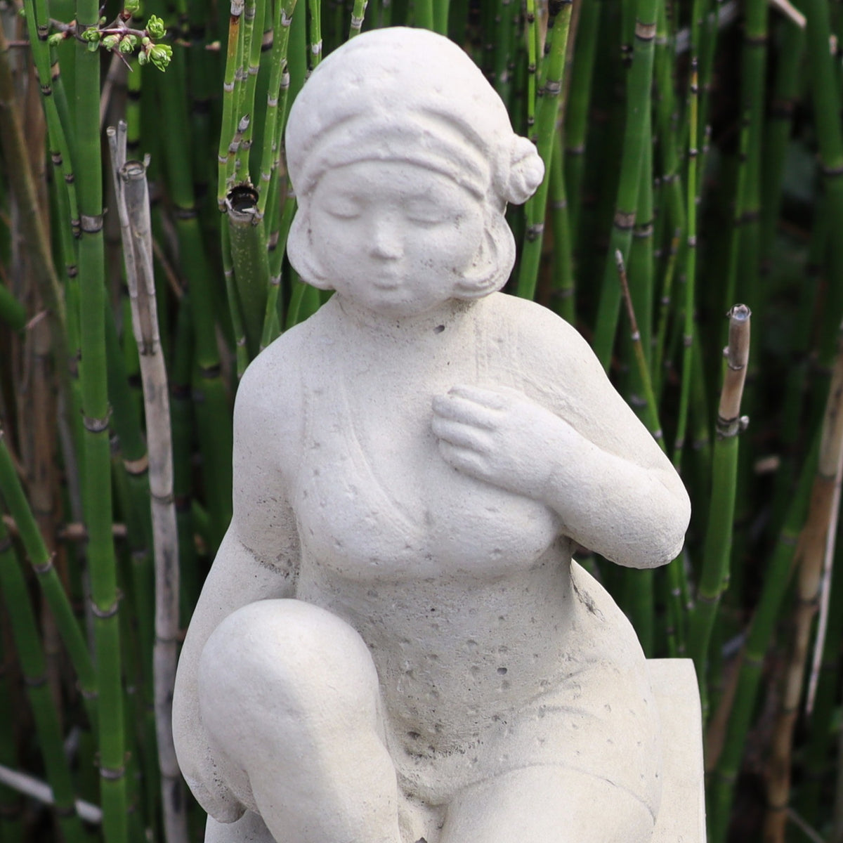 Die UV-beständige Gartenfigur "Badenixe" zeigt eine weiße Steinfigur einer sitzenden Frau mit Kopftuch, einer Hand auf der Brust und einer auf dem Knie - ideal als Gartendekoration vor hohen Bambusstauden.