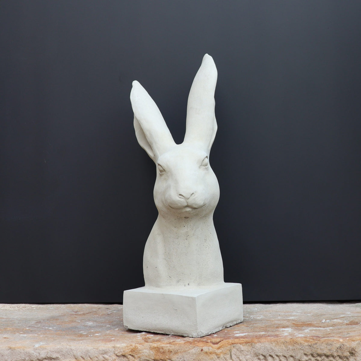 Die handgefertigte Steinfigur "Hase" aus Deutschland zeigt den weißen Kopf und Oberkörper eines Kaninchens auf quadratischem Sockel, arrangiert auf grobem Stein mit dunklem Hintergrund.