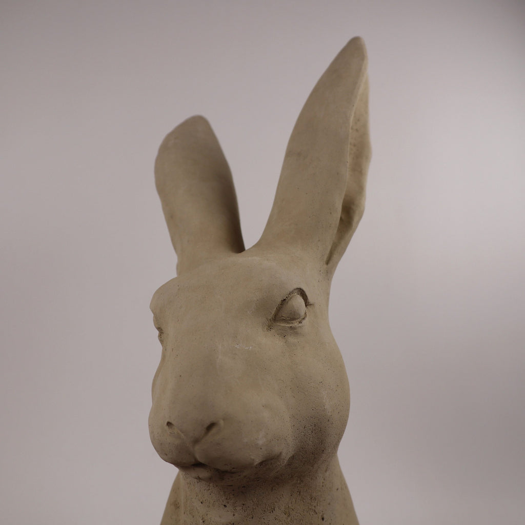 Eine Nahaufnahme der handgefertigten Steinfigur "Hase" aus Deutschland, einer beigen Kaninchenskulptur mit großen Ohren, strukturierter Oberfläche und neutralem Ausdruck vor einem schlichten hellen Hintergrund.