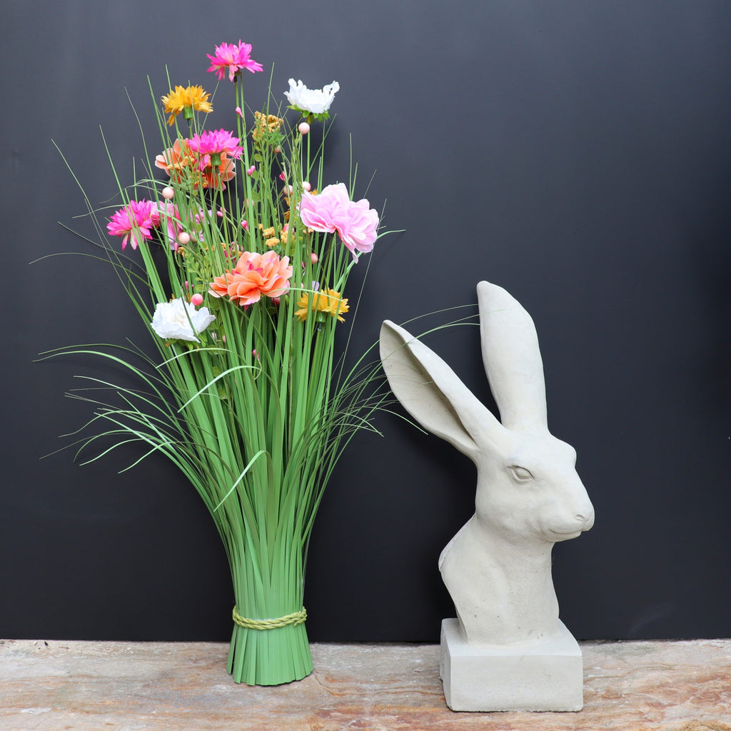 Die handgefertigte Steinfigur "Hase" aus Deutschland mit langen Ohren steht neben einem Strauß bunter Kunstblumen im hohen Gras auf Stein, vor schwarzem Hintergrund.
