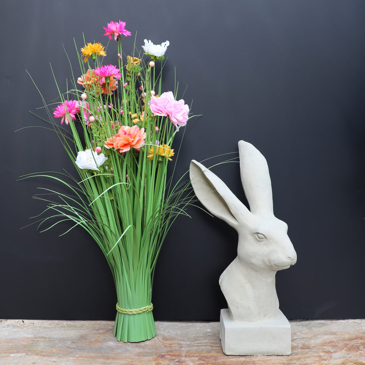 Die handgefertigte Steinfigur "Hase" aus Deutschland mit langen Ohren steht neben einem Strauß bunter Kunstblumen im hohen Gras auf Stein, vor schwarzem Hintergrund.