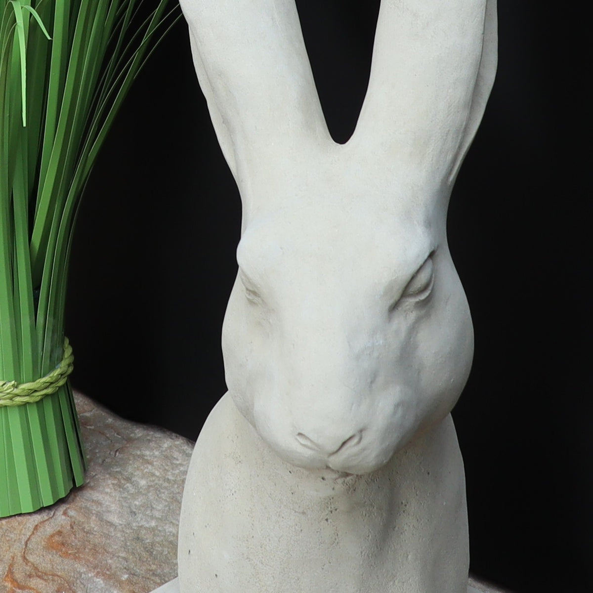 Die handgefertigte Steinfigur "Hase" aus Deutschland, eine glatte weiße Statue eines Hasen mit aufgerichteten Ohren, sitzt auf Marmor. Links im Hintergrund steht ein grünes Bündel langes Gras, mit Strohband gebunden.