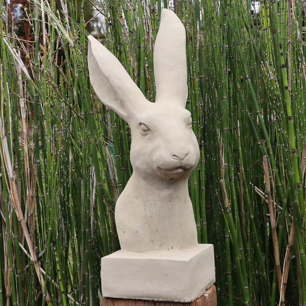 Die handgefertigte Steinfigur "Hase" aus Deutschland zeigt einen Hasenkopf mit langen Ohren und ruhigem Ausdruck auf quadratischem Sockel, dekorativ vor grünen, bambusähnlichen Pflanzen platziert.