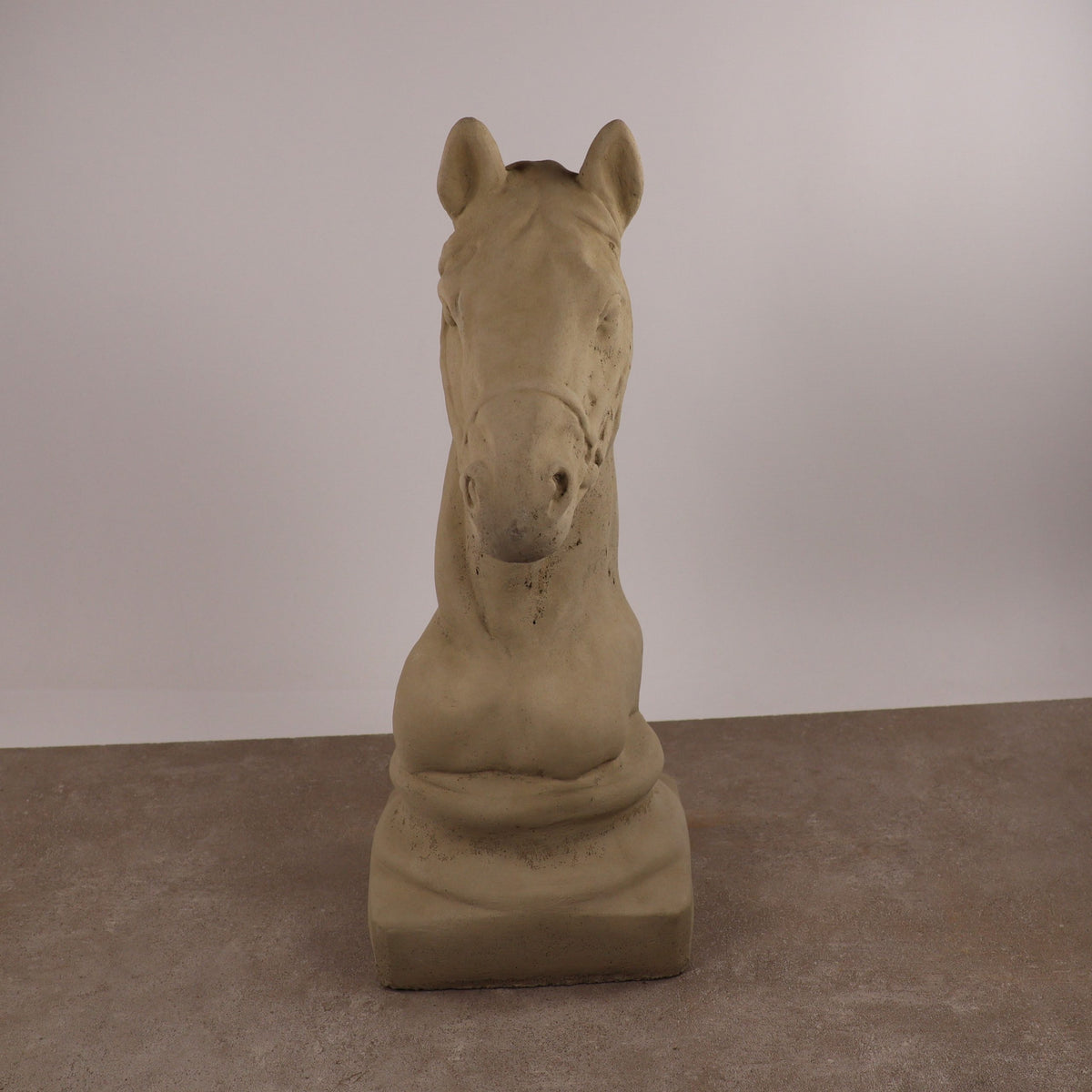 Die "Winterfeste Figur für Outdoor 'Pferd klein'" ist in beigem Stein oder Gips auf einer braunen, strukturierten Oberfläche vor einem hellen Hintergrund dargestellt. Diese Pferdekopfskulptur hat minimale Details und eine glatte, elegante Oberfläche.