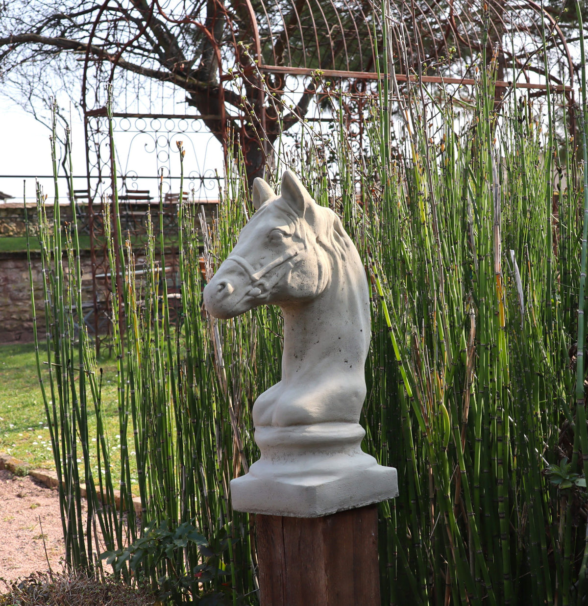 Die Winterfeste Figur für Outdoor "Pferd klein" steht auf einem hölzernen Sockel inmitten von hohem grünen Gras und Gartenstrukturen im Freien, hervorgehoben unter einem teilweise bewölkten Himmel.