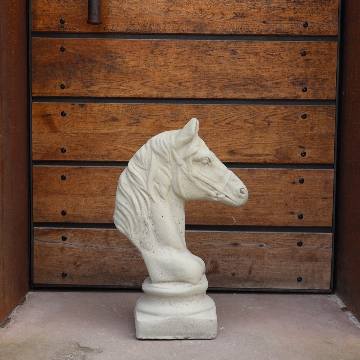 Eine helle "Winterfeste Figur für Outdoor 'Pferd klein'" steht auf einem Betonboden vor einer Holztür mit horizontalen Paneelen und sichtbaren schwarzen Riegeln.