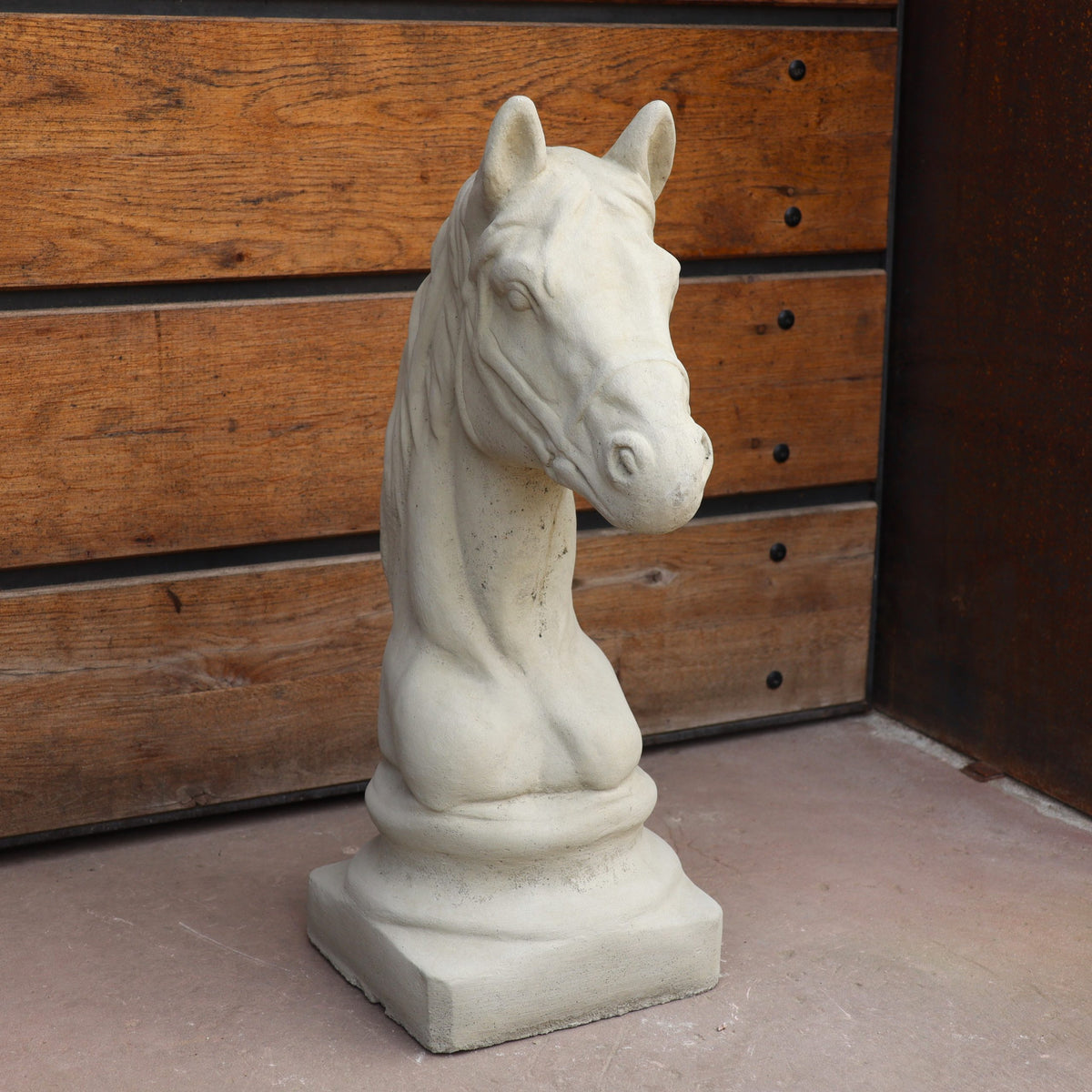 Die Winterfeste Figur für Outdoor "Pferd klein", eine Pferdebüste auf rechteckigem Sockel, steht auf Betonboden vor einer Holzwand mit Metallecke.