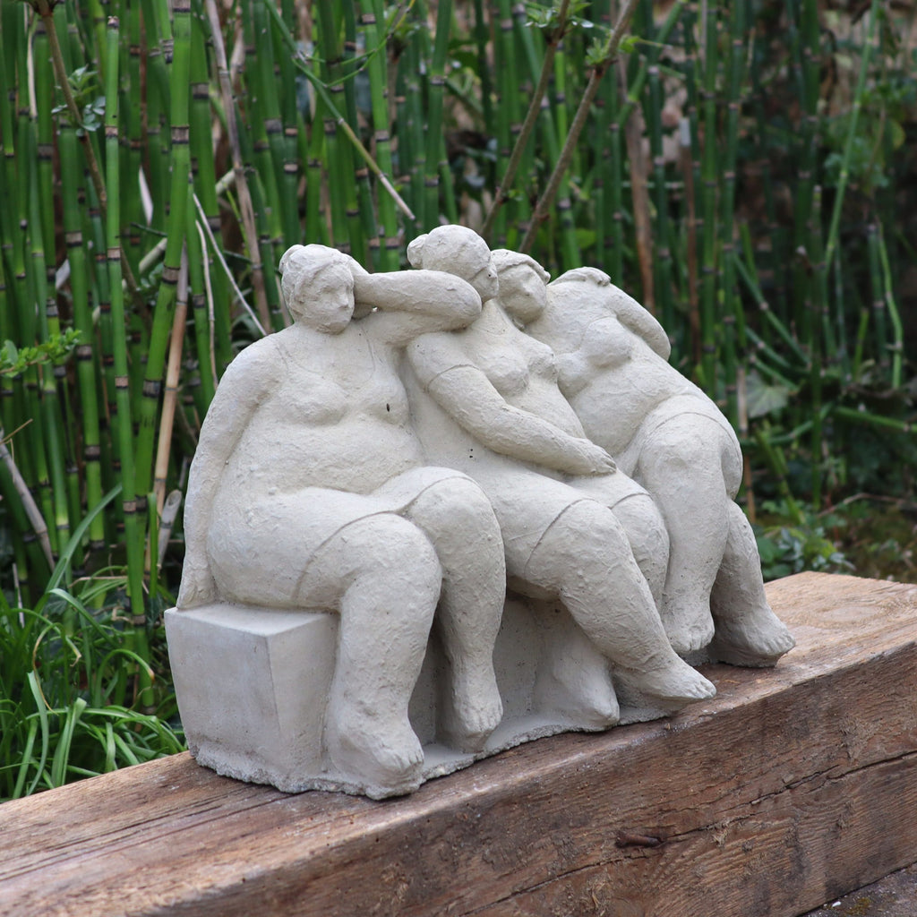 Die charmante Steinfigur "Freundschaft" zeigt drei entspannt sitzende Frauen auf einem Holzstamm, umgeben von grünen Pflanzen - ein handgefertigtes Highlight für Ihren Garten, Made in Germany.