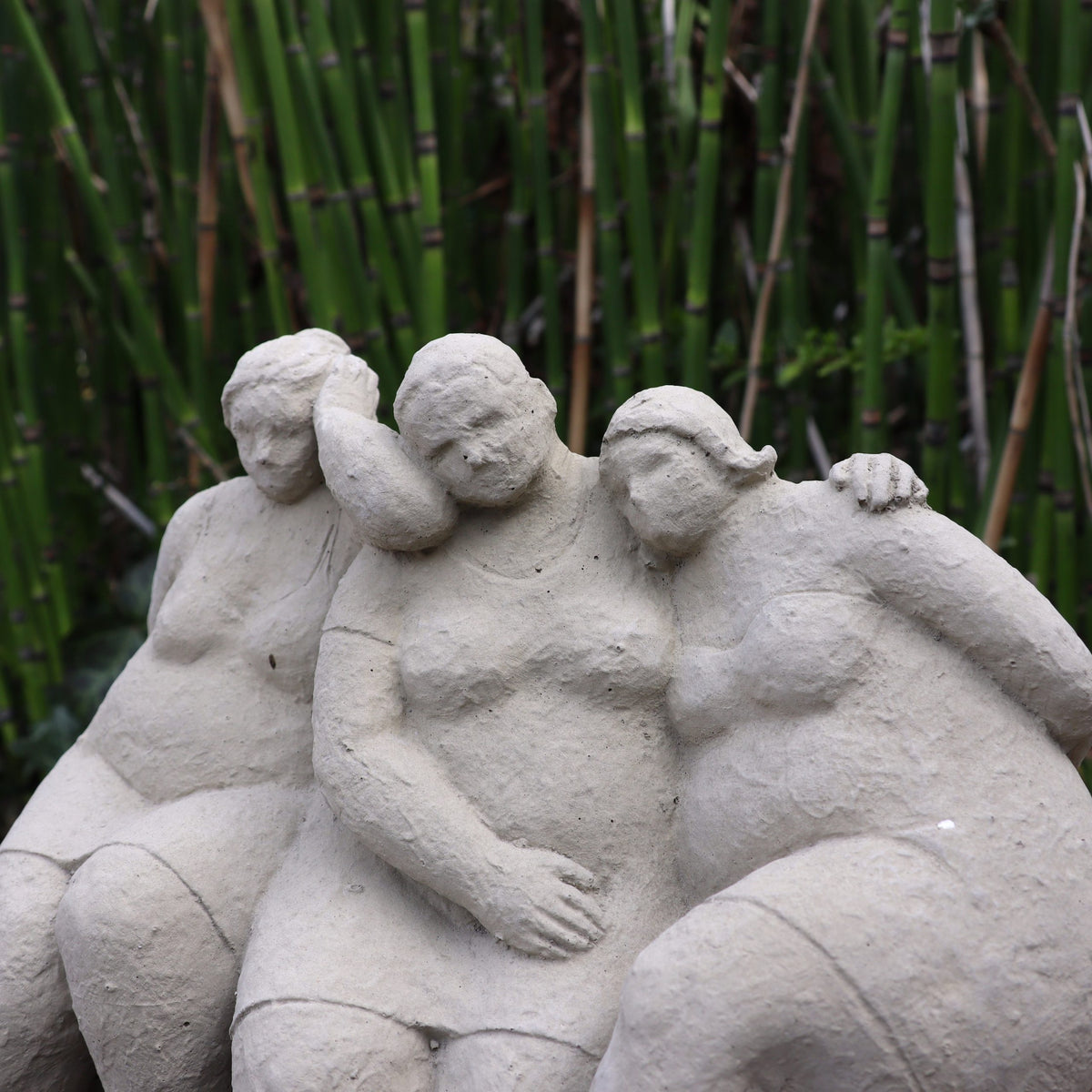 Die charmante Steinfigur "Freundschaft" zeigt drei sitzende Frauen in sanfter Geste, liebevoll handgefertigt in Deutschland - ein Symbol für Gartenliebe vor grünem Bambus als perfekte Deko für Ihren Garten.