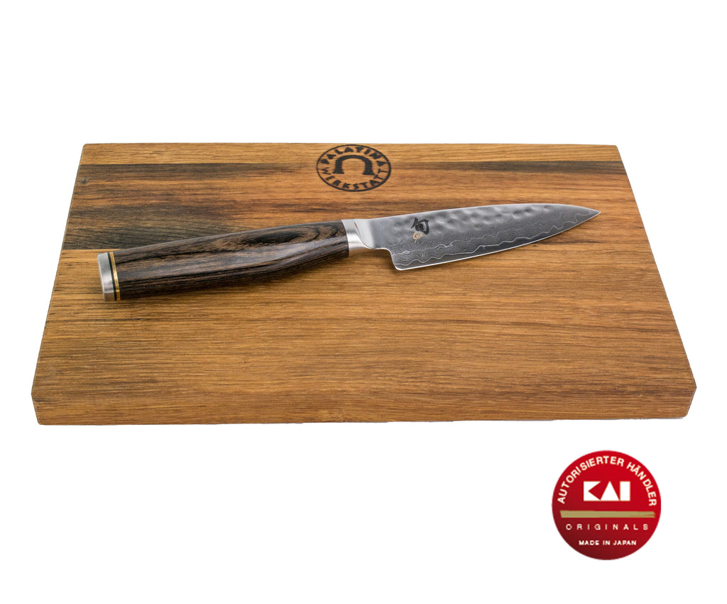 Ein Kai Shun Premier Tim Mälzer Serie TDM-1700 Officemesser mit Holzgriff liegt auf einem handgefertigten Vesperbrett aus alten Fassdauben, 25x15 cm groß. Das Messer hat ein Damaszenermuster auf der Klinge und in der unteren rechten Ecke des Schneidebretts befindet sich ein rotes, rundes Emblem mit deutschem Text, der die Echtheit anzeigt.