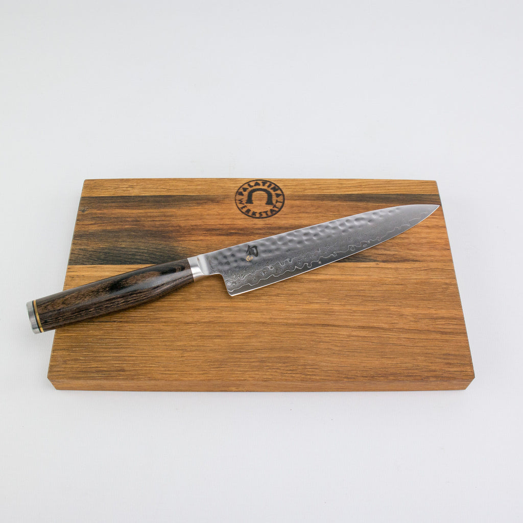 Ein großes Kochmesser aus rostfreiem Stahl mit dunklem Holzgriff, wahrscheinlich das Kai Shun Tim Mälzer Angebotsset Allzweckmesser TDM1701, ruht auf einem handgefertigten Schneidebrett aus Fassdauben (Eiche) 25x15 cm. Das Brett hat oben einen dunkleren Streifen und ist mit einem eingeprägten kreisförmigen Logo mit dem Bild eines Berges und dem Text „HANDMADE 100% NATURAL“ versehen.