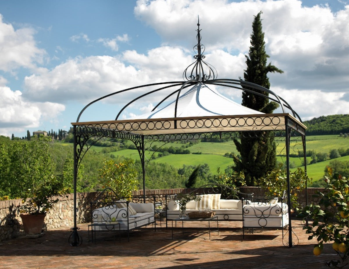 Ein stilvoller Pavillon Tibisco mit schmiedeeisernem Rahmen und elegantem weißen Baldachin steht auf einer geräumigen Backsteinterrasse. Darunter laden gepolsterte Bänke und ein Tibisco-Couchtisch zum gemütlichen Entspannen ein. Im Hintergrund runden sanfte grüne Hügel, hohe Zypressen und ein teilweise bewölkter Himmel die ruhige Szene ab.