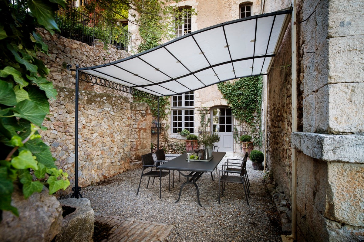 Eine bezaubernde Außenterrasse mit einem schwarzen Metalltisch und Stühlen auf einer Kiesfläche unter einer Anbaupergola Tibisco mit Überdachung aus Polykarbonat für Schatten. Die Umgebung ist von Steinmauern, Topfpflanzen und einer Holztür umgeben, die zu einem mit Efeu bewachsenen Gebäude führt.