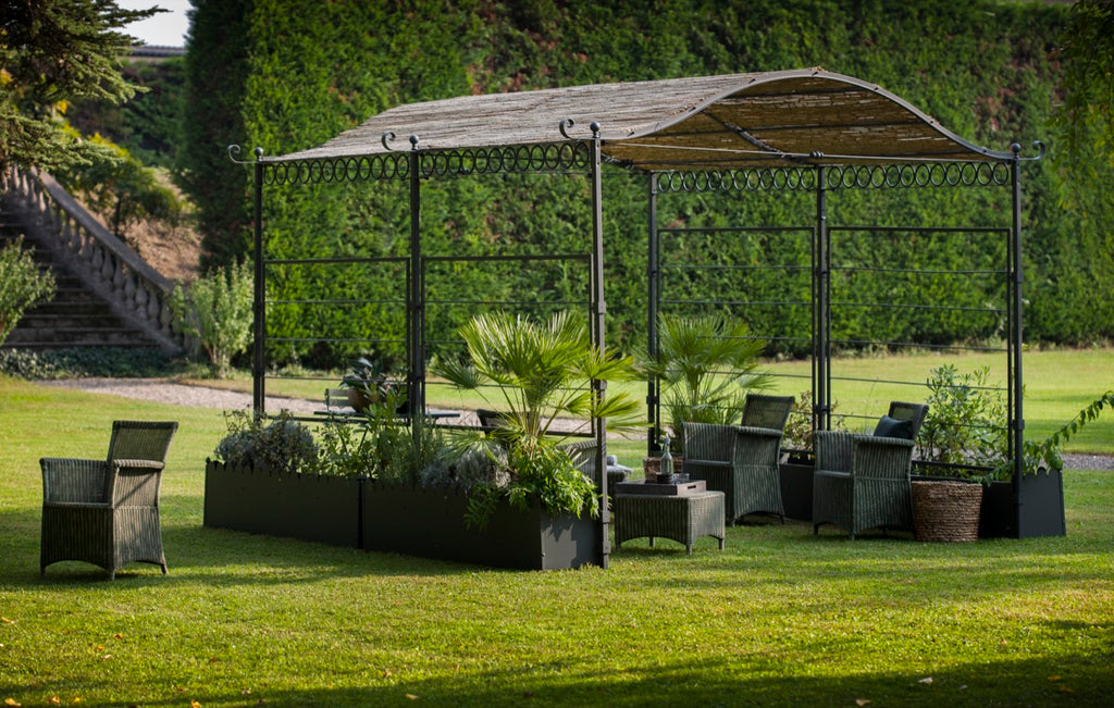 Eine Gartenszene mit der freistehenden Pergola Tibisco mit Überdachung auf einem üppigen grünen Rasen. Im Inneren der Pergola stehen Korbstühle und Topfpflanzen. Dahinter tragen hohe Hecken und Bäume zur ruhigen Kulisse bei. Links sind Stufen zu sehen, die zu einem erhöhten Bereich hinaufführen.