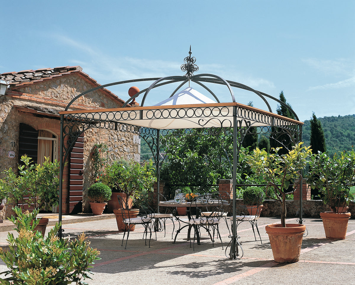 Gezeigt wird eine Terrasse mit einer Pavillon-Tibisco-Struktur, die aufwendig gestaltet ist. Unter dem Pavillon Tibisco stehen ein Metalltisch und mehrere Stühle. Topfpflanzen und Laub umgeben den Bereich, im Hintergrund ein rustikales Steingebäude und eine malerische Aussicht auf Bäume und Hügel.