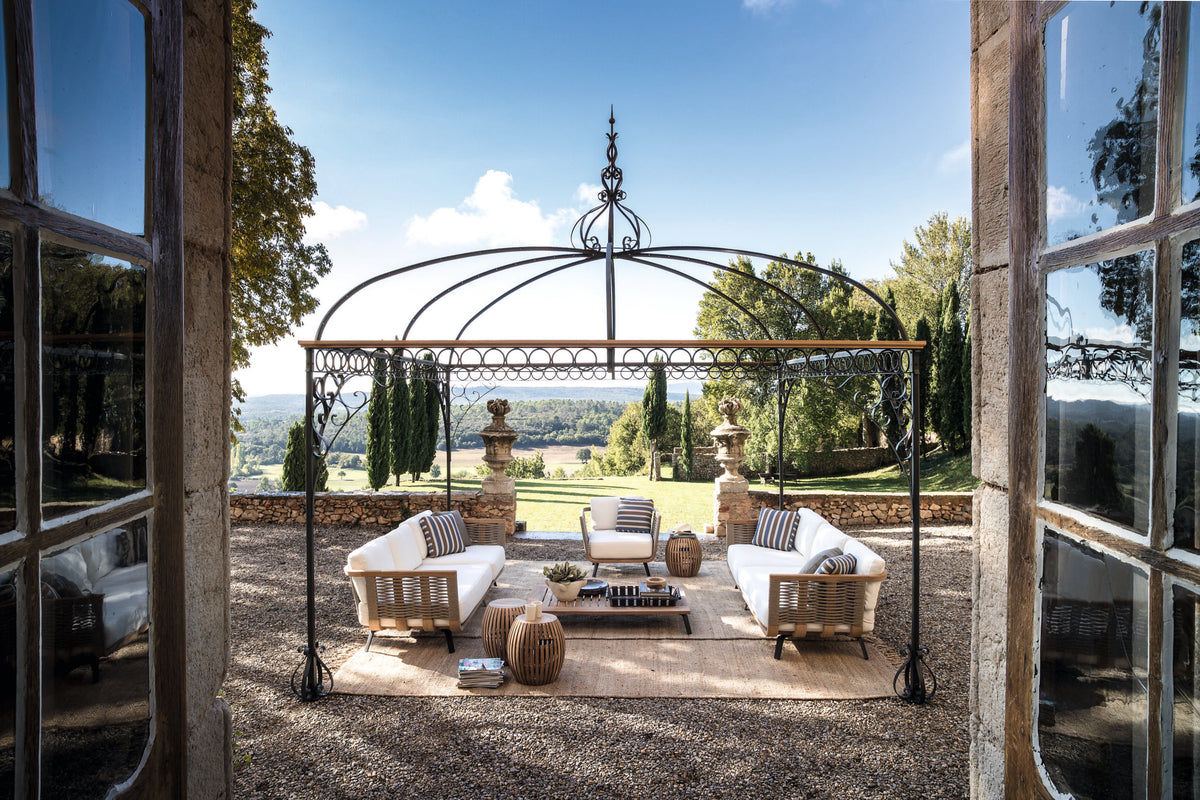 Ein eleganter Sitzbereich im Freien verfügt über weiße Polstermöbel unter einem dekorativen Pavillon Tibisco. Von hier aus hat man einen Blick auf eine malerische Landschaft mit üppigem Grün und fernen Hügeln, eingerahmt von einem offenen Eingang mit großen Glasfenstern.