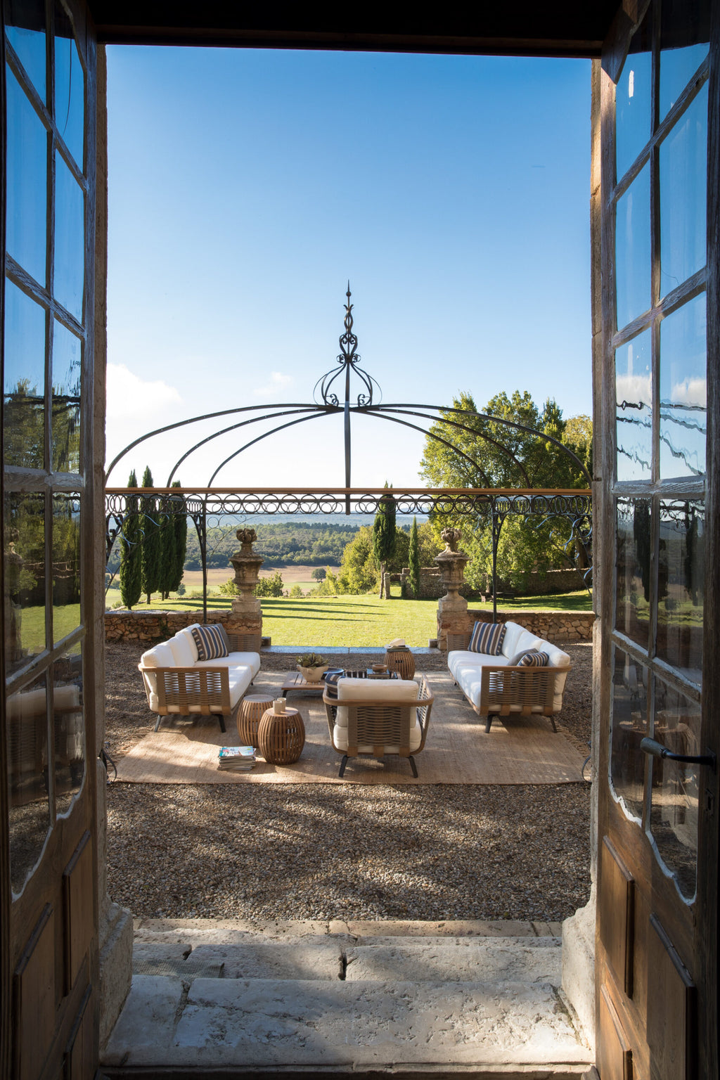 Durch eine offene Tür gelangt man auf eine malerische Terrasse mit eleganten Gartenmöbeln, darunter weiß gepolsterte Stühle und Korbtische, mit Blick auf eine malerische Landschaft mit sanften Hügeln und klarem blauen Himmel, eingerahmt von einem dekorativen Pavillon Tibisco.