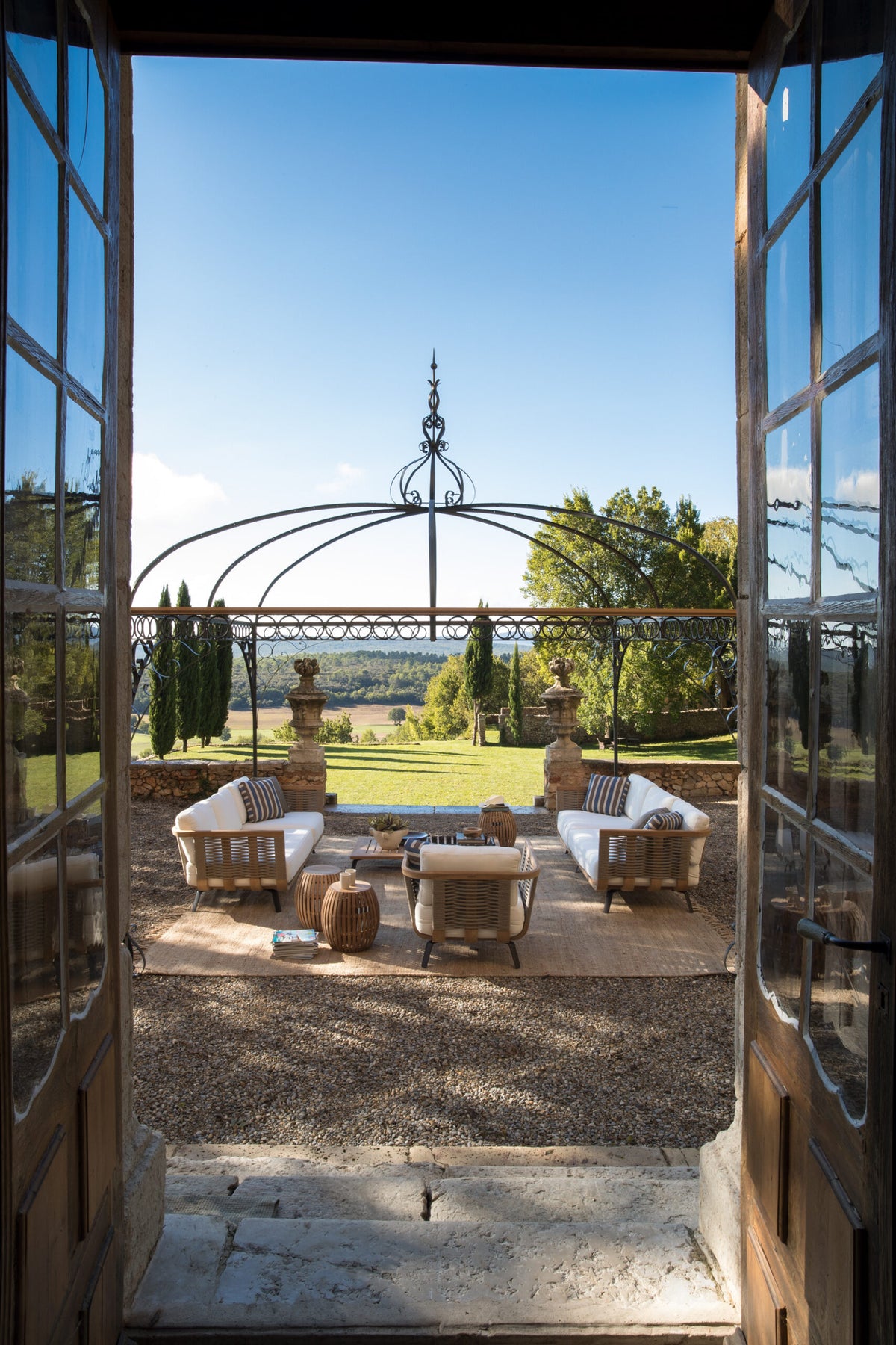 Durch eine offene Tür gelangt man auf eine malerische Terrasse mit eleganten Gartenmöbeln, darunter weiß gepolsterte Stühle und Korbtische, mit Blick auf eine malerische Landschaft mit sanften Hügeln und klarem blauen Himmel, eingerahmt von einem dekorativen Pavillon Tibisco.
