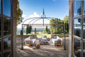 Auf einer geräumigen Außenterrasse steht der Pavillon Tibisco, eine schmiedeeiserne Pergola, unter der ein stilvoller Sitzbereich mit weißen gepolsterten Sofas und Holzmöbeln eingerichtet ist. Der Pavillon bietet Aussicht auf eine weitläufige, malerische Landschaft mit Bäumen, Hügeln und einem klaren blauen Himmel in der Ferne.