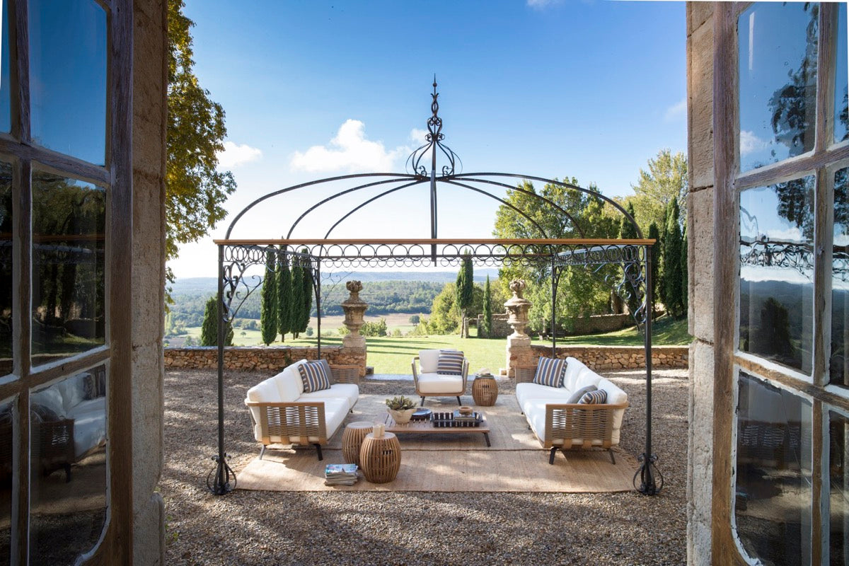 Auf einer geräumigen Außenterrasse steht der Pavillon Tibisco, eine schmiedeeiserne Pergola, unter der ein stilvoller Sitzbereich mit weißen gepolsterten Sofas und Holzmöbeln eingerichtet ist. Der Pavillon bietet Aussicht auf eine weitläufige, malerische Landschaft mit Bäumen, Hügeln und einem klaren blauen Himmel in der Ferne.