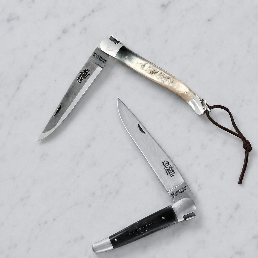 Auf einer hellgrauen Marmorfläche liegen zwei Klappmesser mit unterschiedlichen Griffmaterialien offen; das eine ist ein „Forge de Laguiole“-Taschenmesser (11 cm) aus rot gepresster Verbundfaser und Messing, während das andere einen dunklen Griff mit einer metallischen Spitze aufweist.