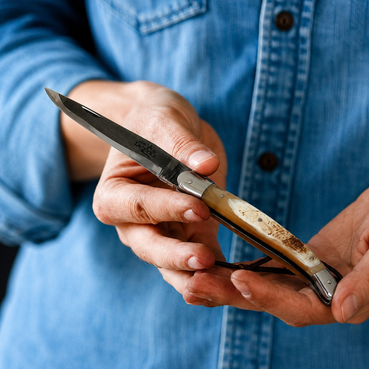 Eine Person in einem blauen Jeanshemd hält ein geöffnetes Klappmesser „Forge de Laguiole Taschenmesser 11 cm“ aus rot gepresstem Verbundfaser- und Messingmaterial in den Händen und zeigt dabei die Klinge und den Griff.
