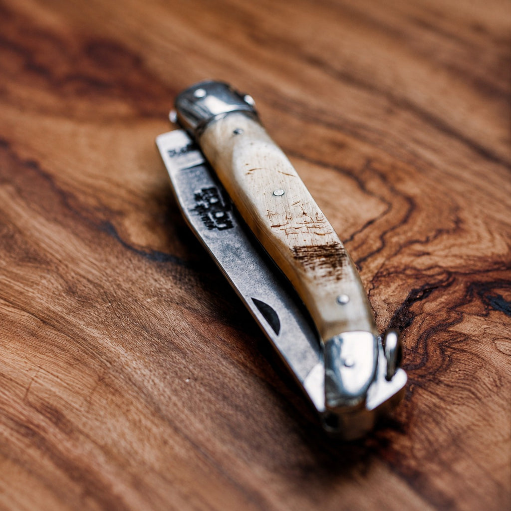 Ein Klappmesser mit einem abgenutzten, hellen Griff liegt geschlossen auf einer strukturierten Holzoberfläche und erinnert an das elegante Design eines „Forge de Laguiole Taschenmessers 11 cm Aubrac Horn, poliert mit Öse“.