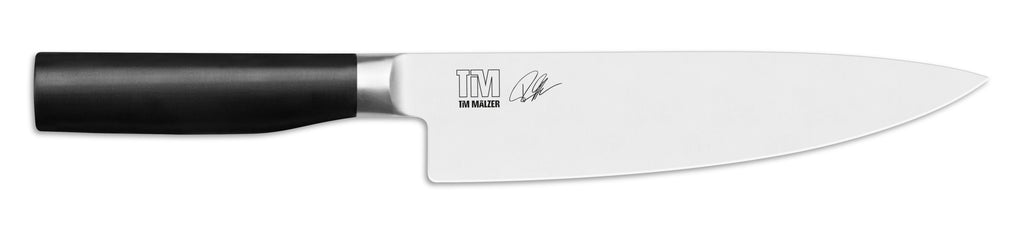 Das KAI X-MAS SELECTION DREAMBOX Tim Mälzer Kamagata Kochmesser hat einen schwarzen Griff und eine glänzende silberne Klinge, die mit dem unverwechselbaren Tim Mälzer Kamagata-Logo mit „TM“ und einer Signatur verziert ist. Dieses exquisite Werkzeug vereint Eleganz mit Funktionalität und ist damit die ideale Wahl für jeden Küchenliebhaber.