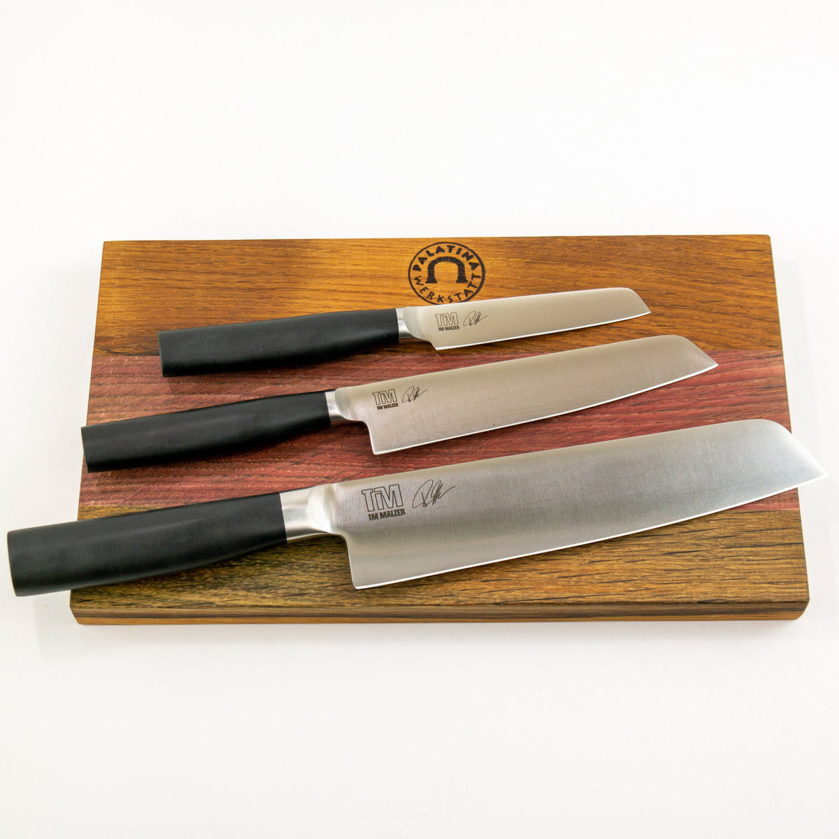 Drei Küchenmesser mit schwarzen Griffen, jeweils in unterschiedlichen Größen, auf einem massiven Holzschneidebrett von 30x18 cm ausgelegt und mit der Aufschrift „PLATINUM SERIES“ beschriftet. Die Klingen sind mit eingravierten Markierungen und Signaturen versehen. Dieses exquisite Kai Shun Tim Mälzer Kamagata Geschenkset ist perfekt für jeden Kochliebhaber.