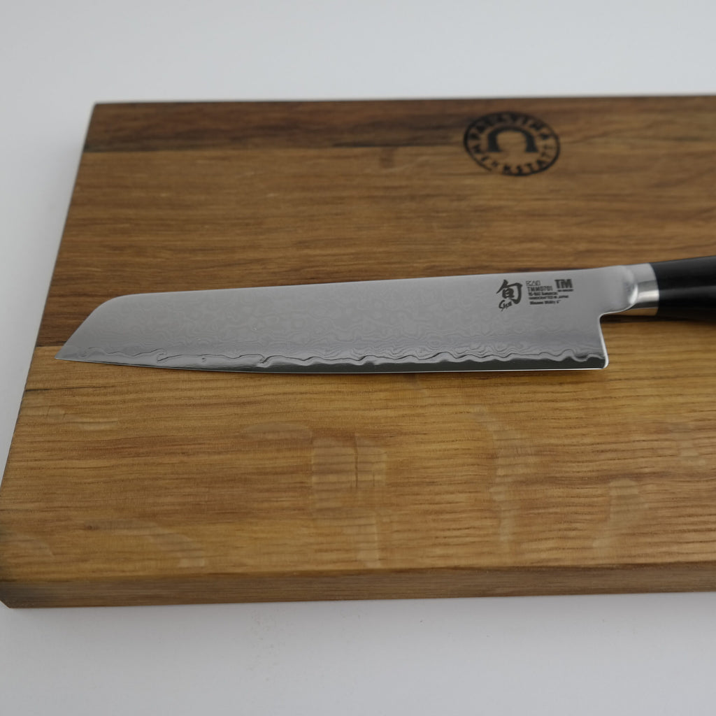 Ein Kai Shun Premier Tim Mälzer Minamo TMM-0701, ein ultrascharfes Allzweckmesser mit 15 cm langer Klinge und dunklem Griff, ruht auf einem Schneidebrett aus altem Fassholz (Eiche) mit den Maßen 25x15 cm. Die Klinge mit Wellenmuster zeigt nach links.