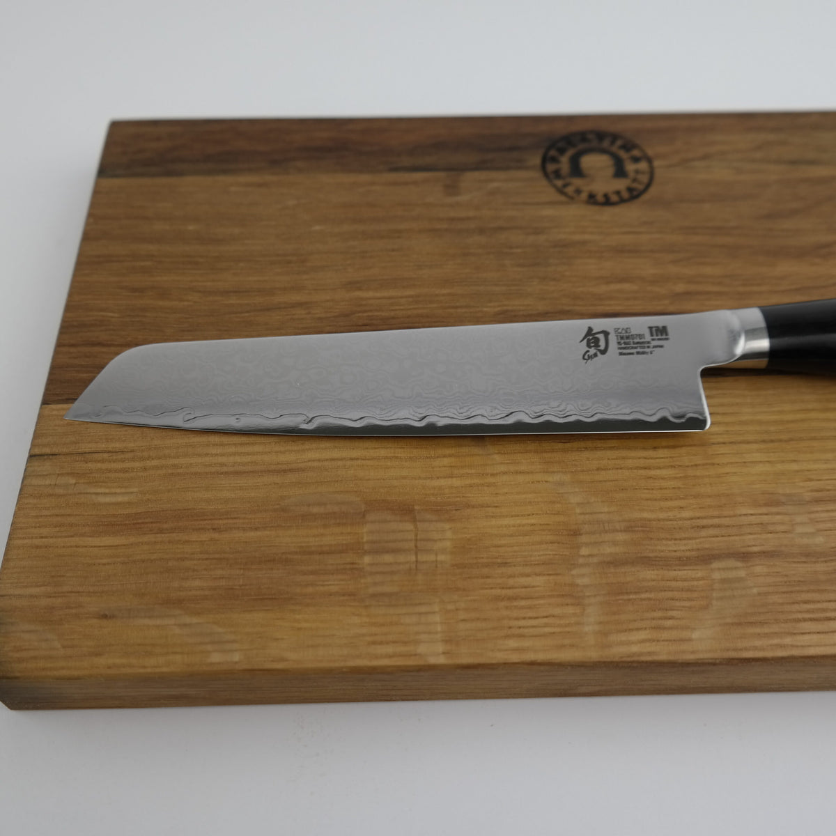 Ein Kai Shun Premier Tim Mälzer Minamo TMM-0701, ein ultrascharfes Allzweckmesser mit 15 cm langer Klinge und dunklem Griff, ruht auf einem Schneidebrett aus altem Fassholz (Eiche) mit den Maßen 25x15 cm. Die Klinge mit Wellenmuster zeigt nach links.